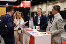 Four individuals stand at an exhibition stand and talk. One person holds a sheet of notes, and promotional materials, badges, and cups are placed on the counter. Other booths and event participants are visible in the background.