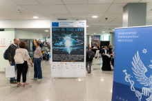 Several people stand and talk in a bright indoor hall next to a large conference poster on a stand. The poster displays the title “ACCORD Interdisciplinary Conference on Drug Sciences” and the slogan “SKY IS THE LIMIT,” along with the location “Warsaw” and the date “May 27–29, 2026.” On the right side, a tall blue banner shows the text “Warszawski Uniwersytet Medyczny” and a white emblem. In the background, more people move through the exhibition space with tables and displays.