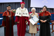  Four people stand on stage. One person holds a document and a case. Behind them, a screen displays “Faculty of Pharmacy WUM 1926–2026”.