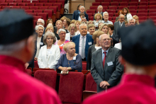 A group of people stands between rows of red seats in an auditorium. The participants have badges and either hold nothing or keep hands folded in front. In the foreground, fragments of figures in red academic gowns are visible on both sides of the frame. In the background, more rows of seating and attendees can be seen.
