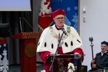 A man in a red academic gown with a rector’s chain and a white fur collar speaks at a lectern with a microphone, resting hands on the podium. In the background there are university symbols and a flag.