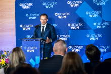 One person stands at a podium with a microphone and speaks to the audience. A backdrop with UCWBK WUM logos is visible behind. The backs of event participants can be seen in the foreground.