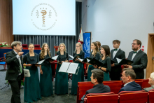 A conductor leads a nine-member choir holding sheet music in a lecture hall, with the Medical University of Warsaw logo displayed behind them.