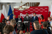 A group of women and men stand in front of a red Perspektywy backdrop during a press event, while journalists and camera operators record the proceedings.