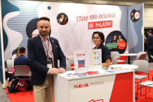 Two individuals stand at a stand marked with the NAWA – Polish National Agency for Academic Exchange logo. Informational materials, badges, and small giveaways are placed on the counter. In the background, there is an exhibition space with meeting areas and a graphic displaying the text “Study and Research in Poland!”.