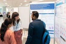 Three people stand in front of scientific poster boards and engage in conversation. One of the poster titles reads “Decoupled Physical and Chemical Stability in Vitamin D3 and D2 Amorphous Solid Dispersions Prepared by Spray Drying and Ball Milling,” with visible sections such as “Objectives” and “Results,” including charts and diagrams. The space is a conference poster area with multiple boards arranged in rows and other attendees in the background.