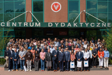 A large group of people stands outside a building for a group photo. Above the entrance, large letters spell “CENTRUM DYDAKTYCZNE,” and above that is a circular emblem with an eagle and surrounding text. Many individuals wear badges on lanyards, and some hold white bags. The building façade features green-framed windows and a red-brown exterior, and the group is arranged on steps in front of the entrance.