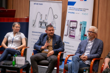Three people sit on a stage in armchairs during a panel discussion. One person in the center holds a handheld microphone and speaks. On a small table in front of them are bottles of water, a glass, and a nameplate. Behind them, large standing banners display laboratory equipment and company names, including “METTLER TOLEDO” and “CEMIS-TECH.”