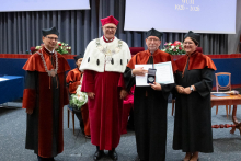  Four people stand on stage. One person holds a document and a case. Behind them, a screen displays “Faculty of Pharmacy WUM 1926–2026”.