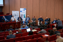 A panel discussion takes place in a lecture hall with several people seated in a row of chairs on stage. One person speaks into a microphone while others listen. In front of the stage, an audience sits in red auditorium seats, and a video camera on a tripod records the event. Two tall banners behind the panel show images of laboratory equipment and company branding.