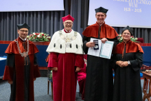  Four people stand on stage. One person holds a document and a case. Behind them, a screen displays “Faculty of Pharmacy WUM 1926–2026”.