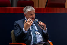 One person sits in an armchair and speaks into a handheld microphone while holding a pen and a document. A conference badge on a lanyard hangs around the neck. The background shows wooden panels and part of the lecture hall stage.