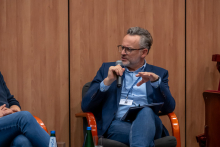 One person sits in an armchair on stage and holds a handheld microphone in one hand and a notepad on their lap. The person gestures with the other hand while speaking. A bottle of water and a glass stand on a small table nearby. The background consists of wooden wall panels.