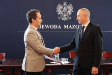 Dwaj mężczyźni stoją naprzeciw siebie przed tłem z napisem „Wojewoda Mazowiecki”. Mężczyźni podają sobie ręce, a ten po lewej trzyma czerwoną teczkę.
