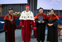  Four people stand on stage. One person holds a document and a case. Behind them, a screen displays “Faculty of Pharmacy WUM 1926–2026”.