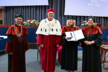 Four people stand on stage. One person holds a document and a case. Behind them, a screen displays “Faculty of Pharmacy WUM 1926–2026”.