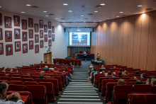 A wide view shows a lecture hall with rows of red auditorium seats arranged in tiers leading down to a stage. On the left wall, numerous framed black-and-white portraits are arranged in a grid. At the front, a large projection screen displays a graphic with the text “SKY IS THE LIMIT,” showing a bright abstract light pattern. Several people sit scattered throughout the seats, some using laptops or looking toward the stage. Near the front, a few individuals stand and move around the lectern and desk area. 