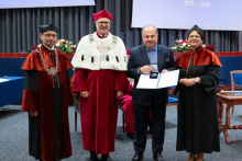 Four people stand on stage. One person holds a document and a case. Behind them, a screen displays “Faculty of Pharmacy WUM 1926–2026”.