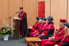A person in a red and black academic gown and cap stands at a lectern holding sheets of paper. Next to the lectern, several seated people are visible, with hands resting on laps and not holding objects. A microphone is placed at the lectern, and a vase with colorful flowers stands nearby. In the background, there is a banner reading “Warszawski Uniwersytet Medyczny Wydział Lekarski.”