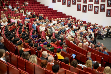 Women and men are seated in an auditorium with red chairs, with some attendees wearing academic gowns and caps. Some participants are holding phones or documents, and framed portraits are displayed on the wall.