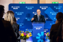 One person stands at a podium and speaks, holding one hand raised in a gesture. Flower arrangements are placed beside the podium. A backdrop with UCWBK WUM logos is visible, and participants stand in the foreground.