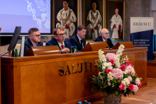 Four people sit at the main table during a conference. Microphones and documents are on the table, and a presentation screen and a KRAUM banner are visible in the background.