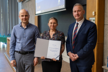 Two men and a woman stand together in a lecture hall, the woman holds an open diploma in her hands.