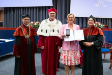 Four people stand on stage. One person holds a document and a case. Behind them, a screen displays “Faculty of Pharmacy WUM 1926–2026”.