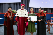 Four people stand on stage. One person holds a document and a case. Behind them, a screen displays “Faculty of Pharmacy WUM 1926–2026”.
