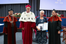 Four people stand on stage. One person holds a document and a case. Behind them, a screen displays “Faculty of Pharmacy WUM 1926–2026”.