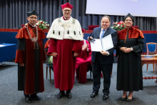  Four people stand on stage. One person holds a document and a case. Behind them, a screen displays “Faculty of Pharmacy WUM 1926–2026”.