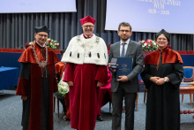  Four people stand on stage. One person holds a document and a case. Behind them, a screen displays “Faculty of Pharmacy WUM 1926–2026”.