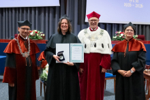  Four people stand on stage. One person holds a document and a case. Behind them, a screen displays “Faculty of Pharmacy WUM 1926–2026”.