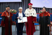  Four people stand on stage. One person holds a document and a case. Behind them, a screen displays “Faculty of Pharmacy WUM 1926–2026”.