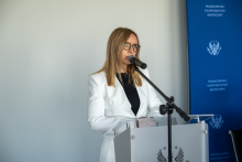 A woman in a white blazer stands at a lectern and delivers a presentation during a conference.
