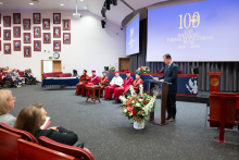A man in a suit speaks at a lectern with a microphone, holding printed speech notes in hands. In the background there are people seated in academic gowns and caps, as well as a screen displaying “100 years of the Faculty of Pharmacy WUM 1926–2026” and floral arrangements.