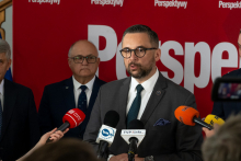 A man in a suit speaks to journalists during a press conference, with microphones held in front of him and other participants standing nearby.
