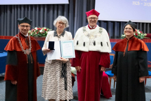  Four people stand on stage. One person holds a document and a case. Behind them, a screen displays “Faculty of Pharmacy WUM 1926–2026”.