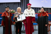  Four people stand on stage. One person holds a document and a case. Behind them, a screen displays “Faculty of Pharmacy WUM 1926–2026”.