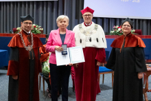  Four people stand on stage. One person holds a document and a case. Behind them, a screen displays “Faculty of Pharmacy WUM 1926–2026”.