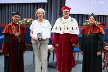  Four people stand on stage. One person holds a document and a case. Behind them, a screen displays “Faculty of Pharmacy WUM 1926–2026”.