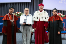 Four people stand on stage. One person holds a document and a case. Behind them, a screen displays “Faculty of Pharmacy WUM 1926–2026”.