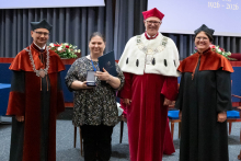  Four people stand on stage. One person holds a document and a case. Behind them, a screen displays “Faculty of Pharmacy WUM 1926–2026”.