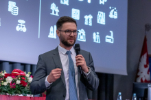 One person stands in front of a screen and speaks into a microphone, giving a presentation. The background shows a screen with icons and elements of a lecture hall.
