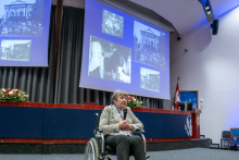 One person in a wheelchair speaks into a microphone in a lecture hall. Behind them, a screen displays black-and-white photographs.