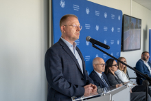 A man wearing glasses and a blue jacket speaks at a lectern, while other conference participants are seated behind him.