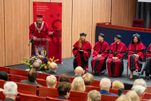 A person in a red academic gown with a decorative chain stands at a lectern and gestures with one hand while speaking. A vase with flowers stands in front of the lectern. Several seated people in gowns keep hands on their laps and do not hold objects. In the foreground, rows of red seats and the audience are visible.