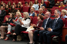 Women and men are seated in red chairs and applauding, with some holding bags or phones in their hands. In the background there is an audience seated across several rows of the auditorium.