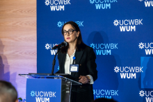 One person stands at a podium and speaks to the event participants. A backdrop with UCWBK WUM logos is visible behind.
