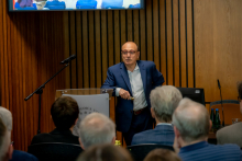 A man stands at a lectern and speaks to an audience, holding a remote control in his hand.